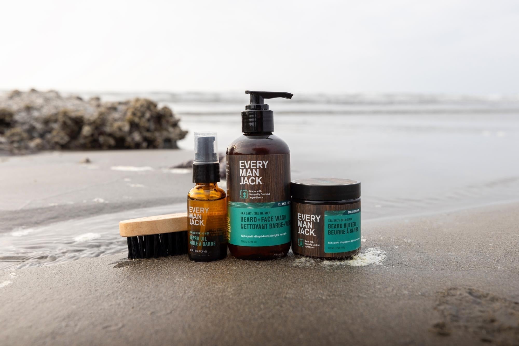 Three Every Man Jack beard grooming products on a beach: beard wash, beard oil, and beard butter.