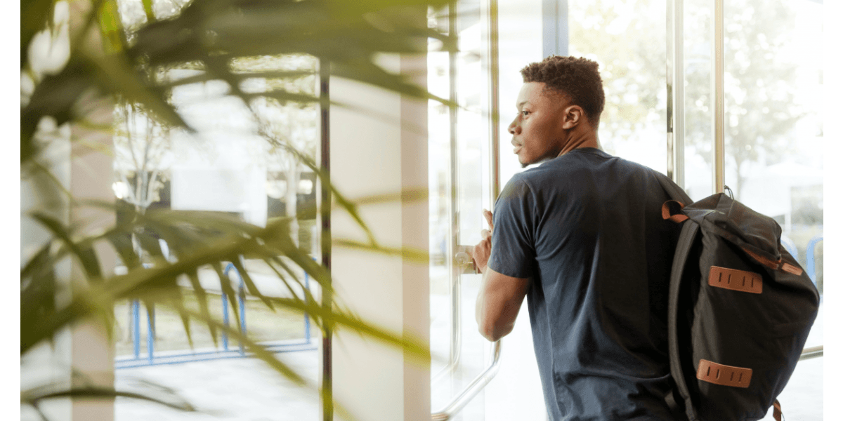 A young man with a backpack looking out a glass door, surrounded by greenery.