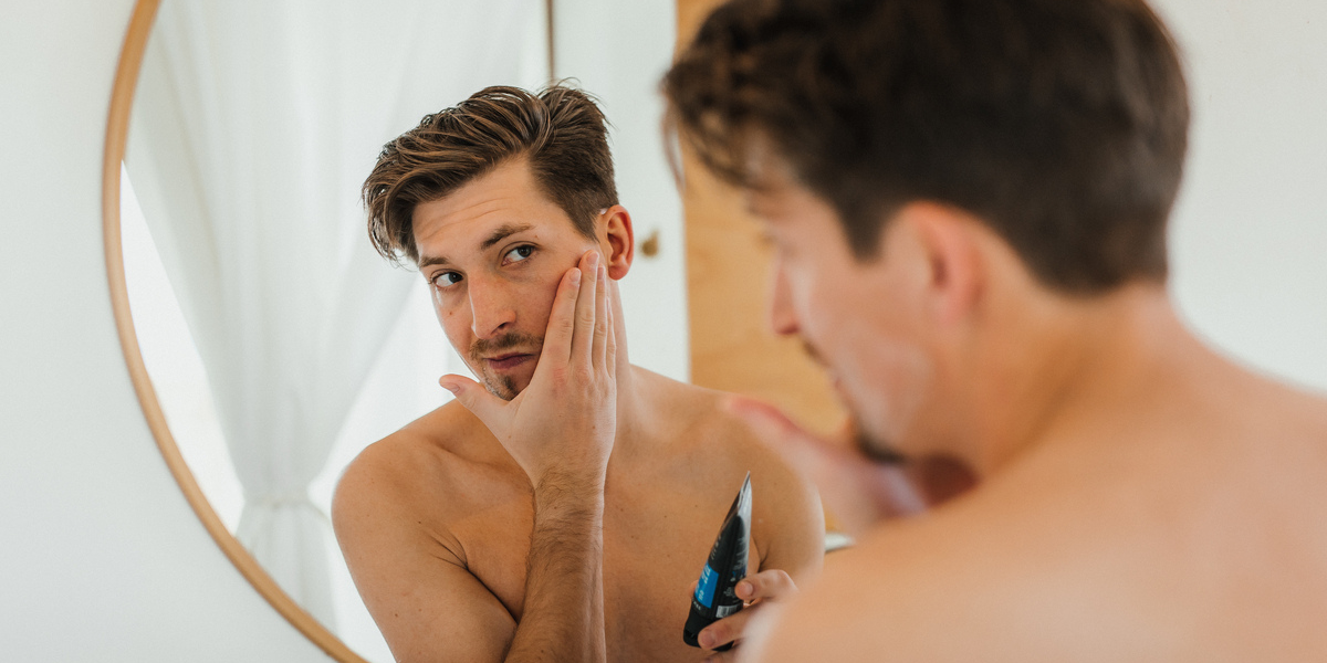 A man applying grooming cream to his face in front of a mirror.