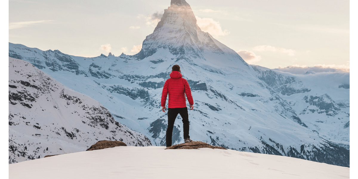 A person in a red jacket standing on a snowy mountain overlooking a peak.