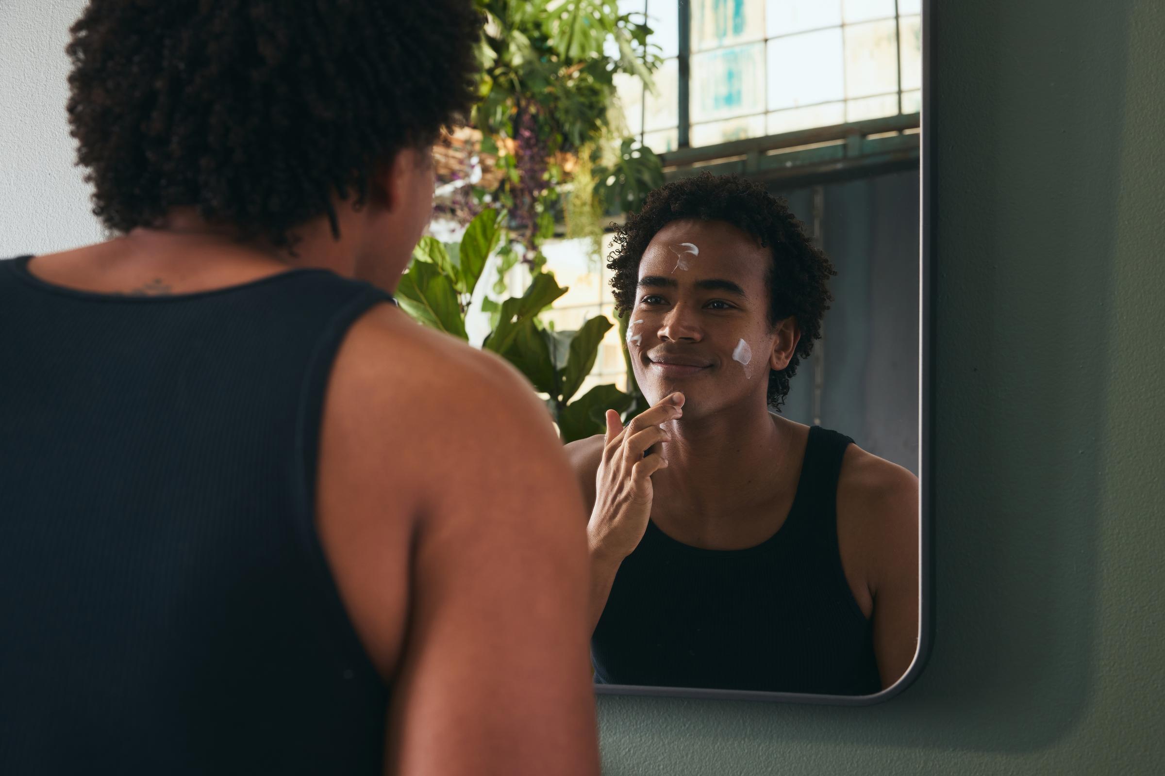 A person applying moisturizer to their face in front of a mirror, surrounded by plants.