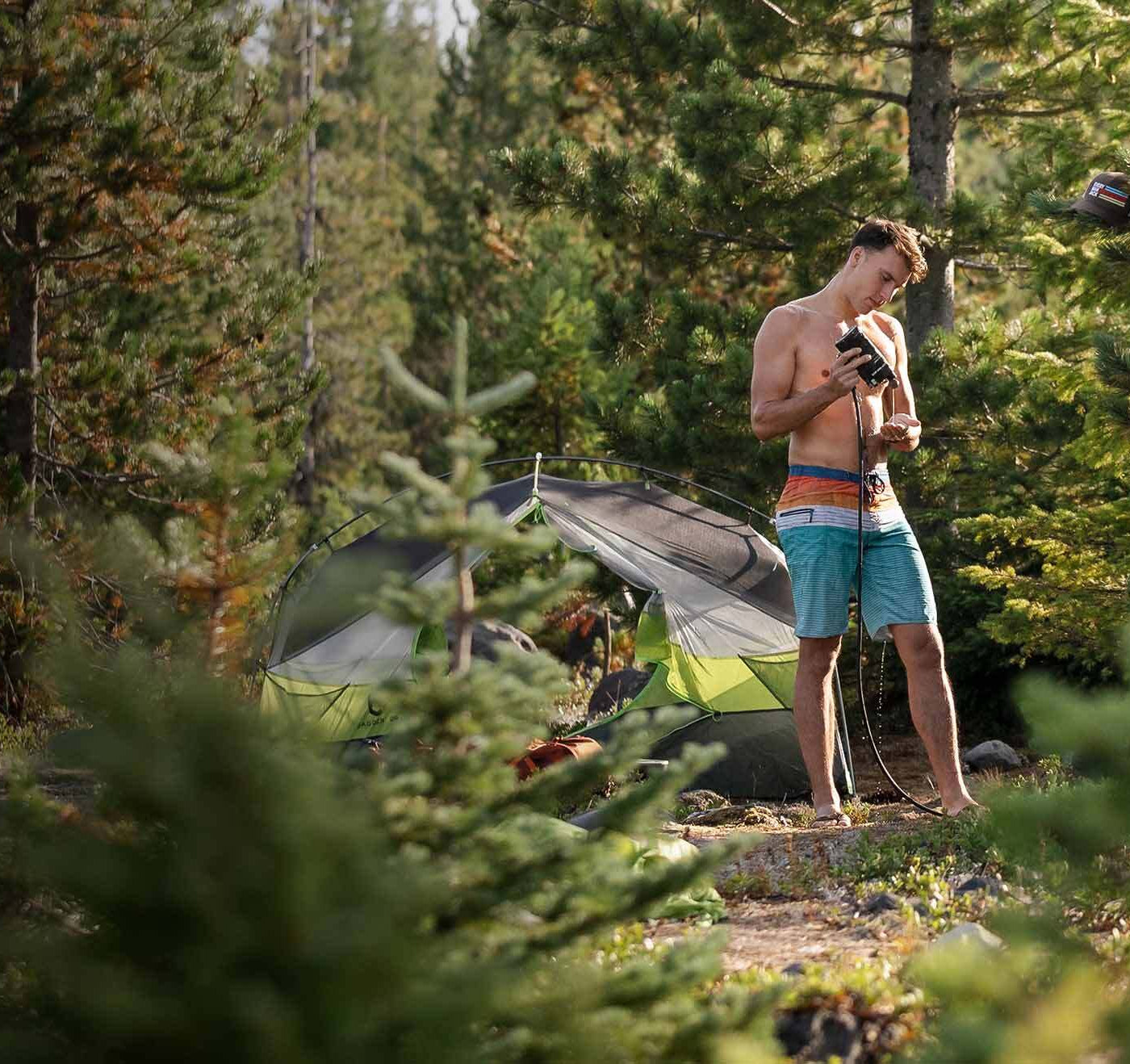 A man in swim trunks holding a grooming product in a forest setting.