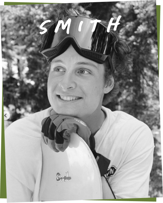 Black and white portrait of a smiling person named Smith, wearing goggles and holding a surfboard.