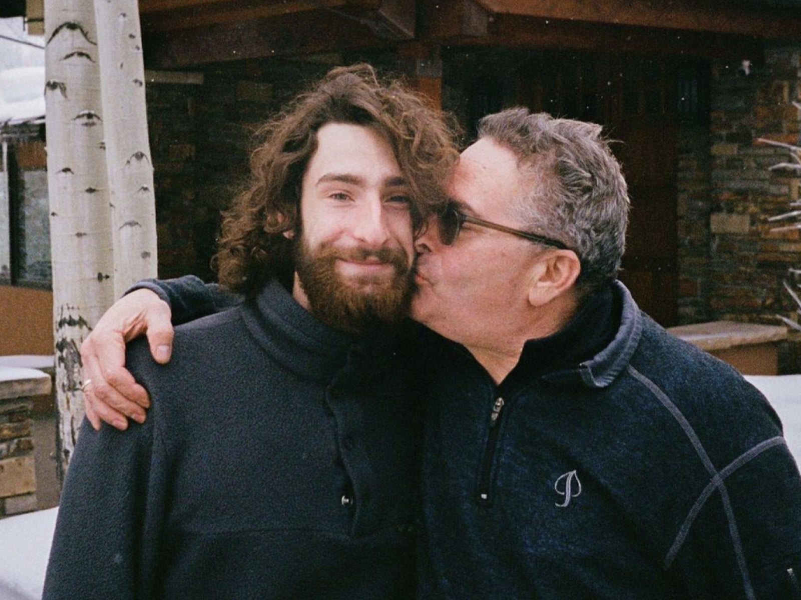 A man kisses another man on the cheek, both smiling, in a snowy outdoor setting.