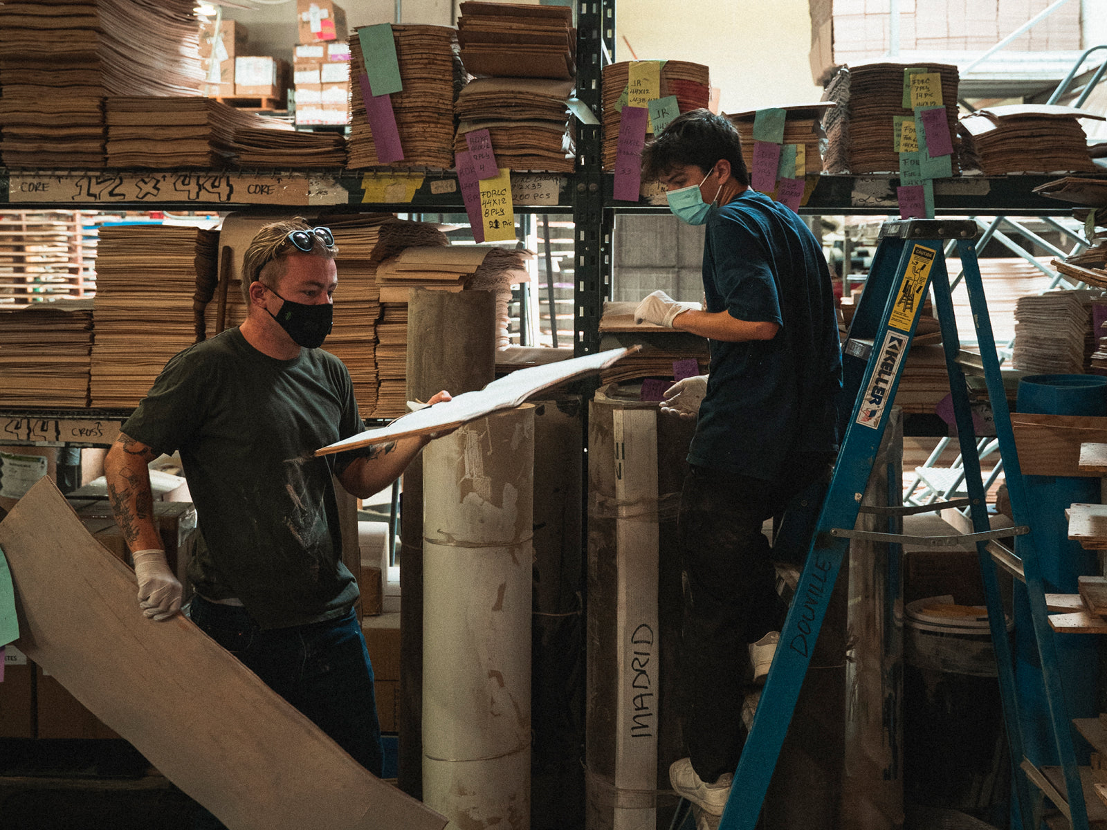 Two individuals in a warehouse examining large sheets of material while wearing masks.