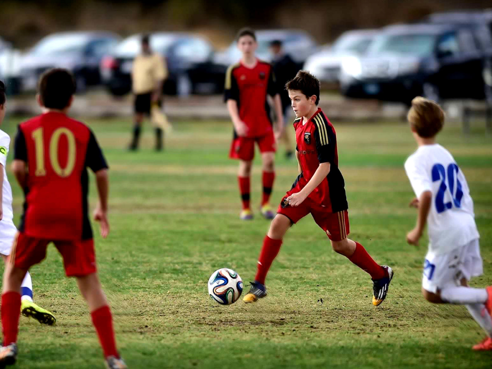 A youth soccer player in a red and black uniform dribbles the ball on a field during a match.