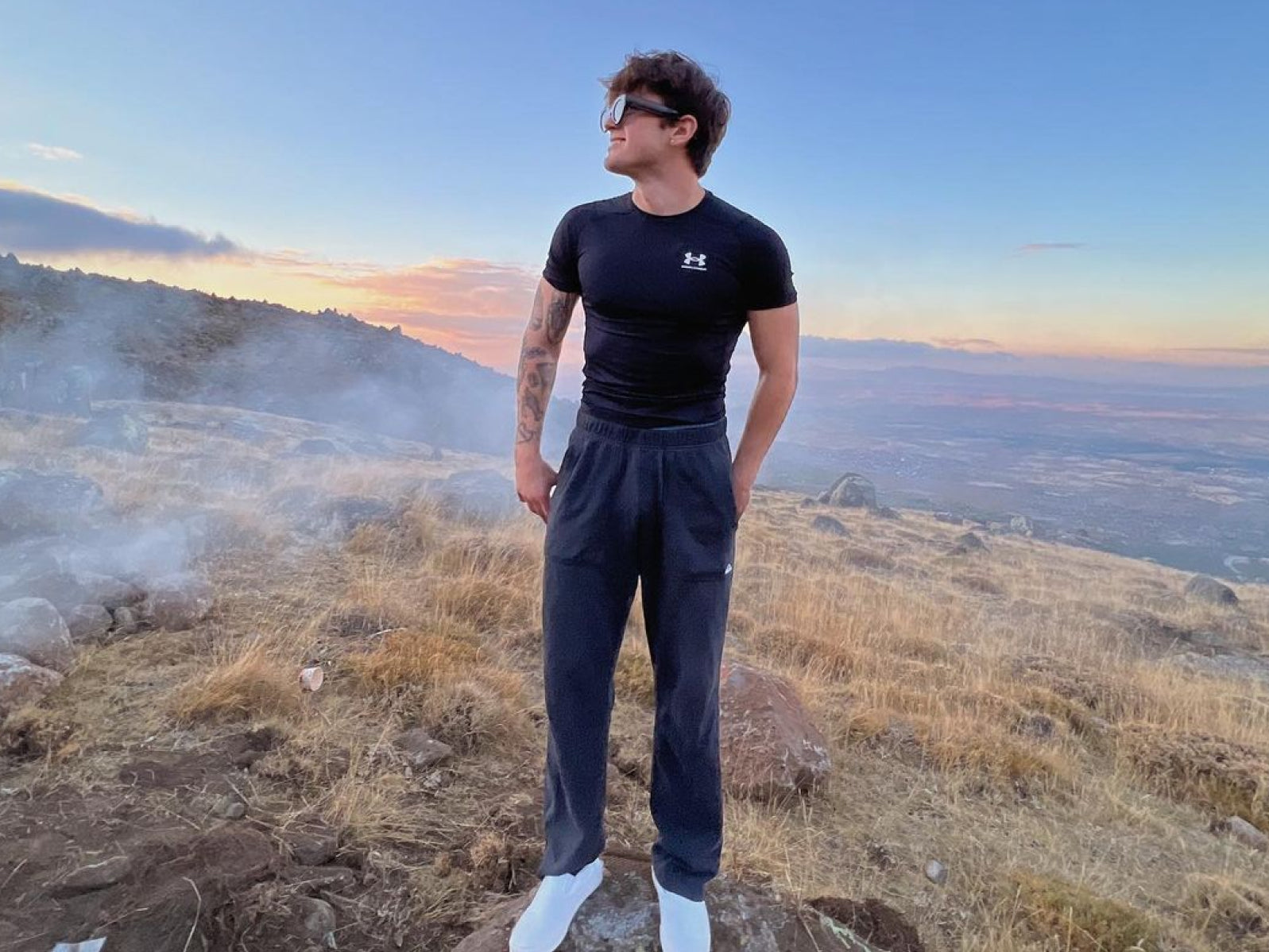 A person in a black athletic shirt and gray pants standing on a rocky hillside during sunset.