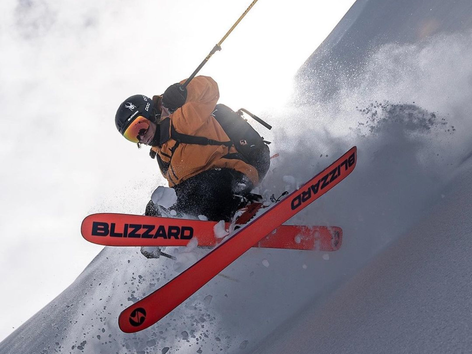 A skier in an orange jacket skiing down a snowy slope on Blizzard skis.