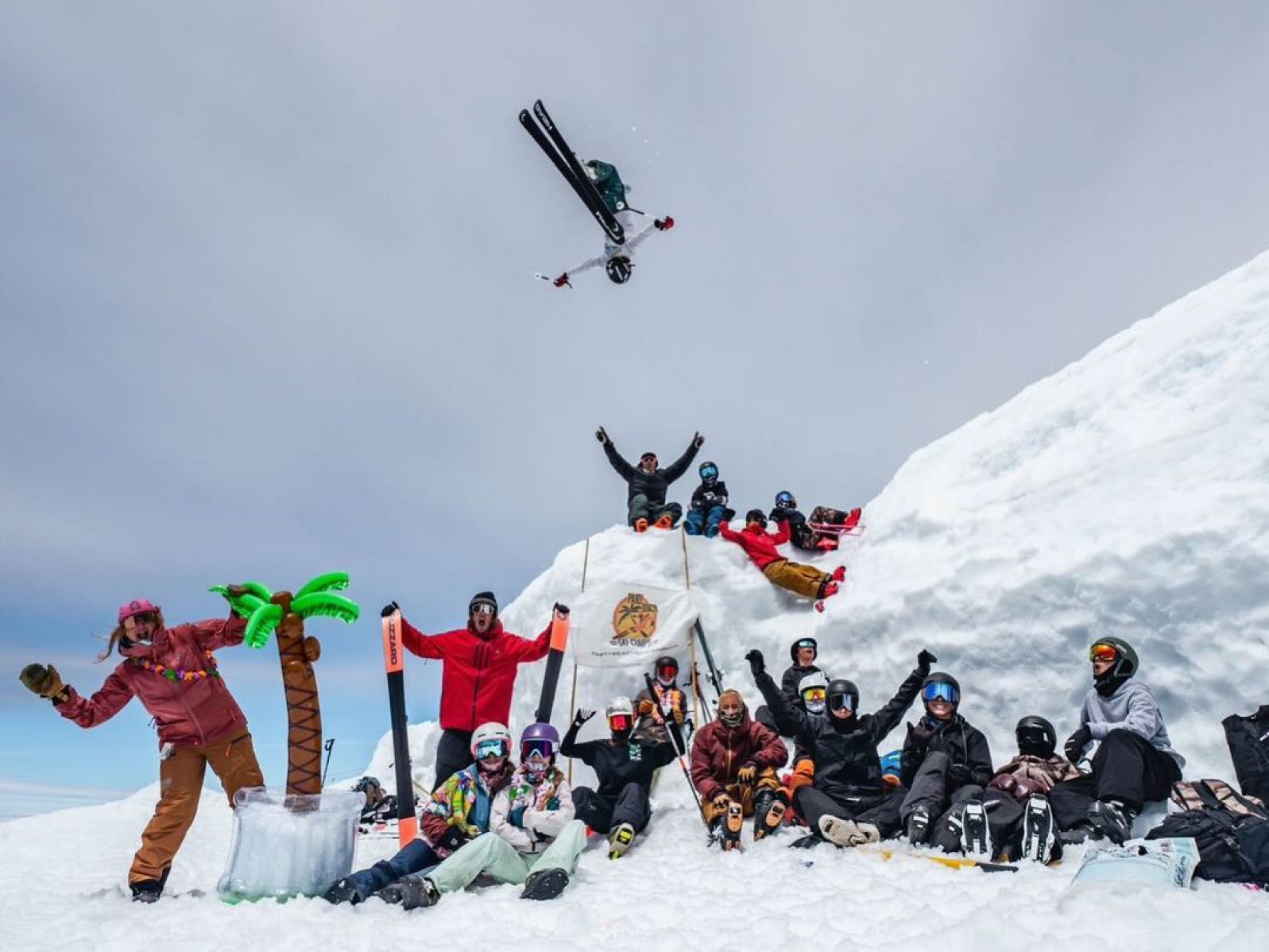 A group of skiers celebrating on a snowy mountain with palm trees and ski gear.
