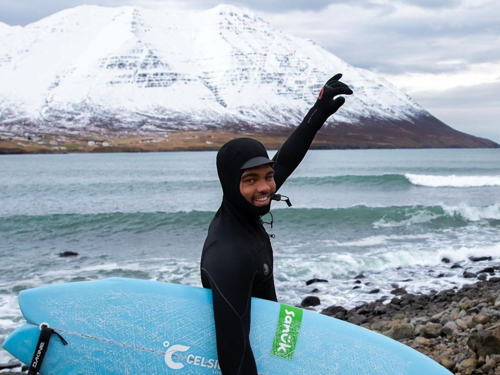 A surfer in a wetsuit holding a blue surfboard, smiling and waving, with snowy mountains and ocean in the background.