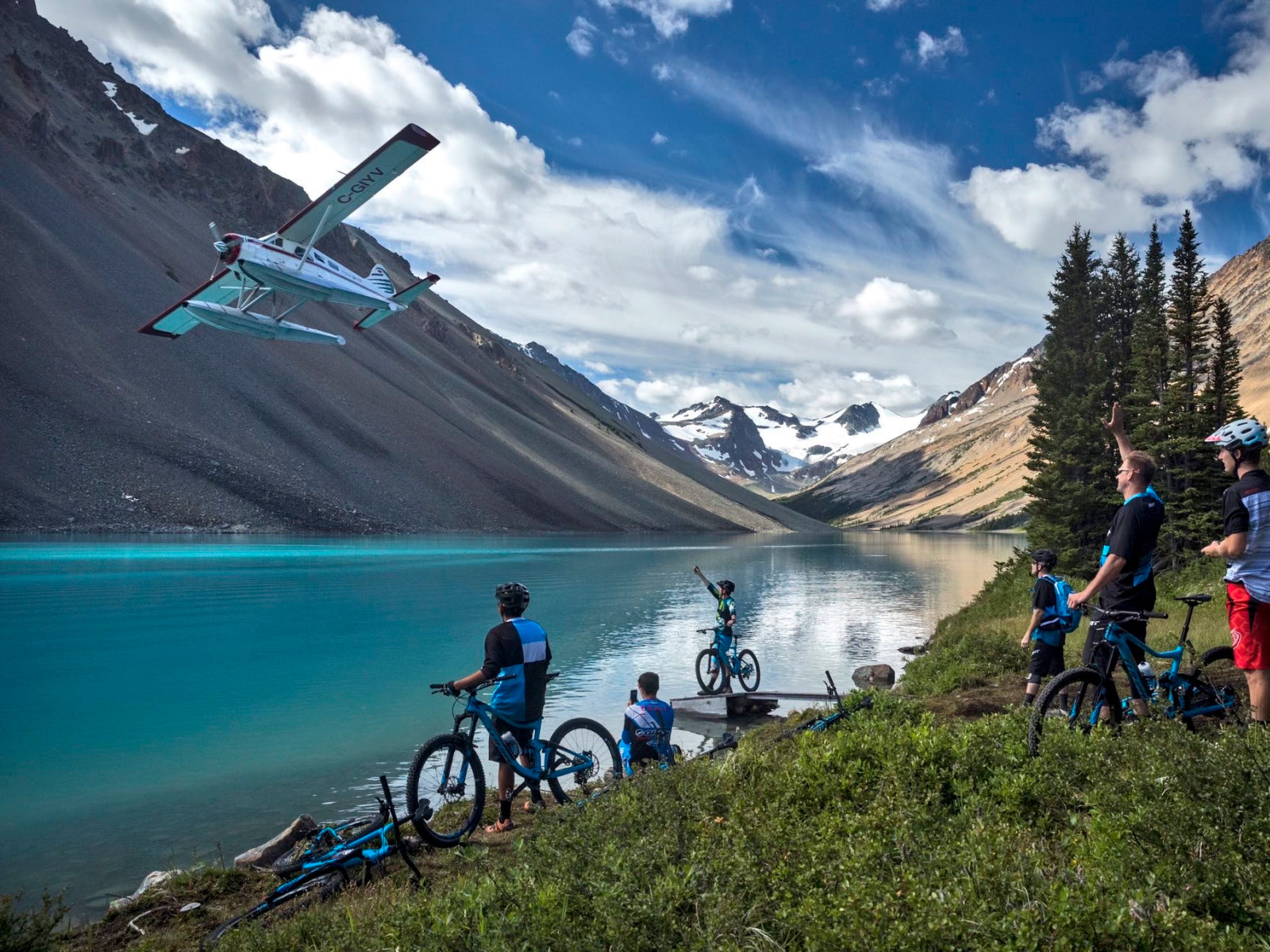 A seaplane flying over a turquoise lake with mountain bikers on the shore and mountains in the background.
