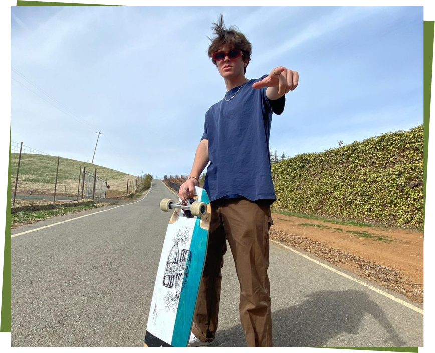 Cole Trotta skateboarding on a road, wearing sunglasses and a blue shirt, pointing forward.