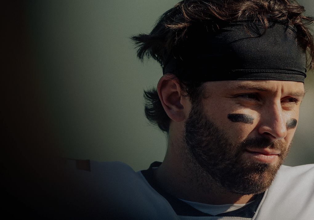 Close-up of a man in a football jersey wearing a headband and eye black, looking to the right against a muted outdoor background