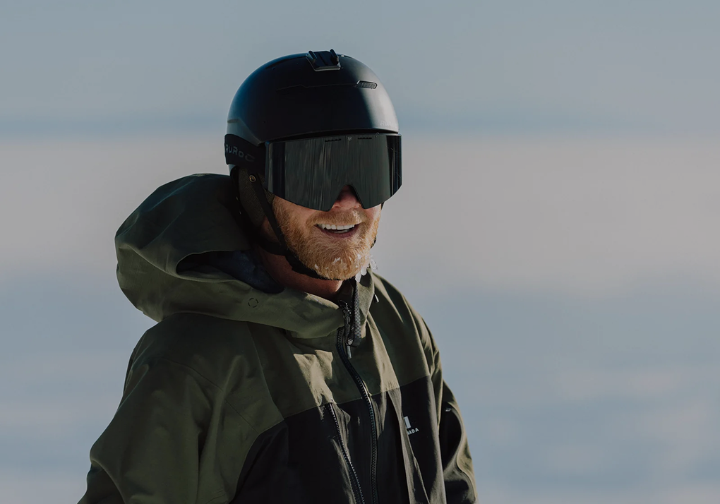 Skier smiling on a snowy slope wearing a black helmet, mirrored goggles, and green jacket