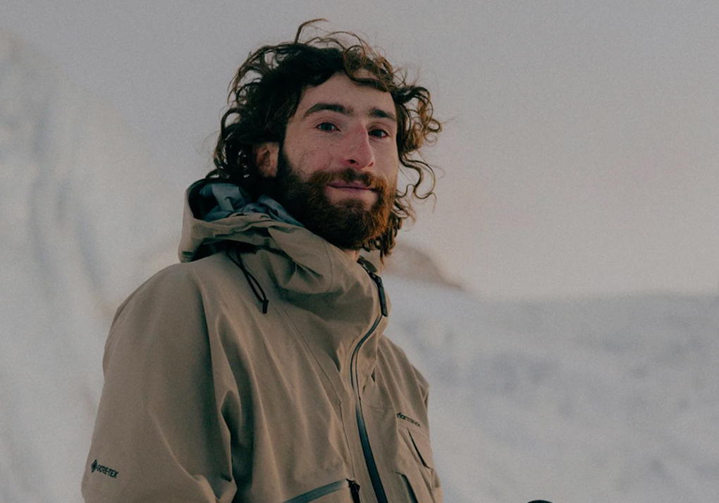 Bearded man standing in a snowy landscape wearing a tan hooded jacket, looking at the camera