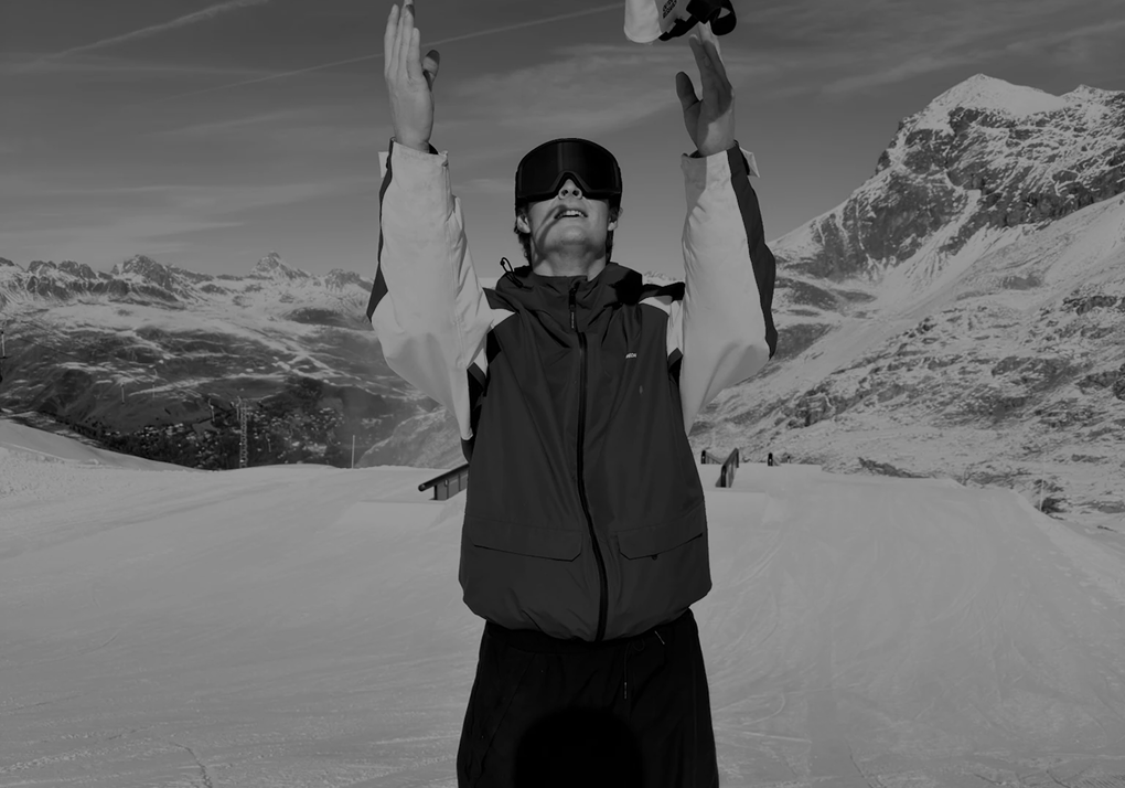 Snowboarder raising his arms to catch a hat on a snow-covered mountain slope with distant peaks