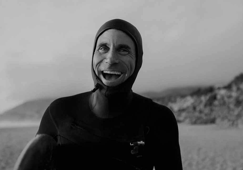 Smiling man in a wetsuit and hood holding a surfboard on a sandy beach with rocky cliffs in the background