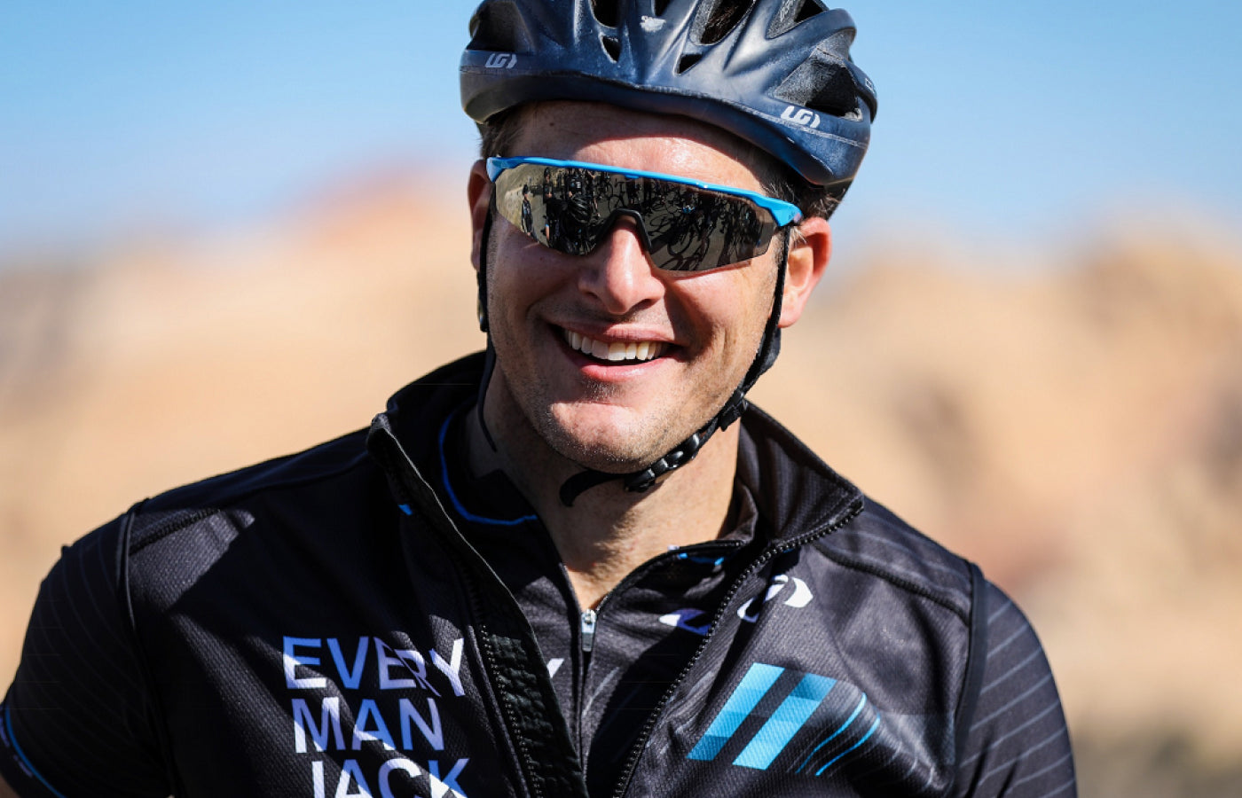 A smiling cyclist in a branded Every Man Jack jersey and sunglasses, outdoors.