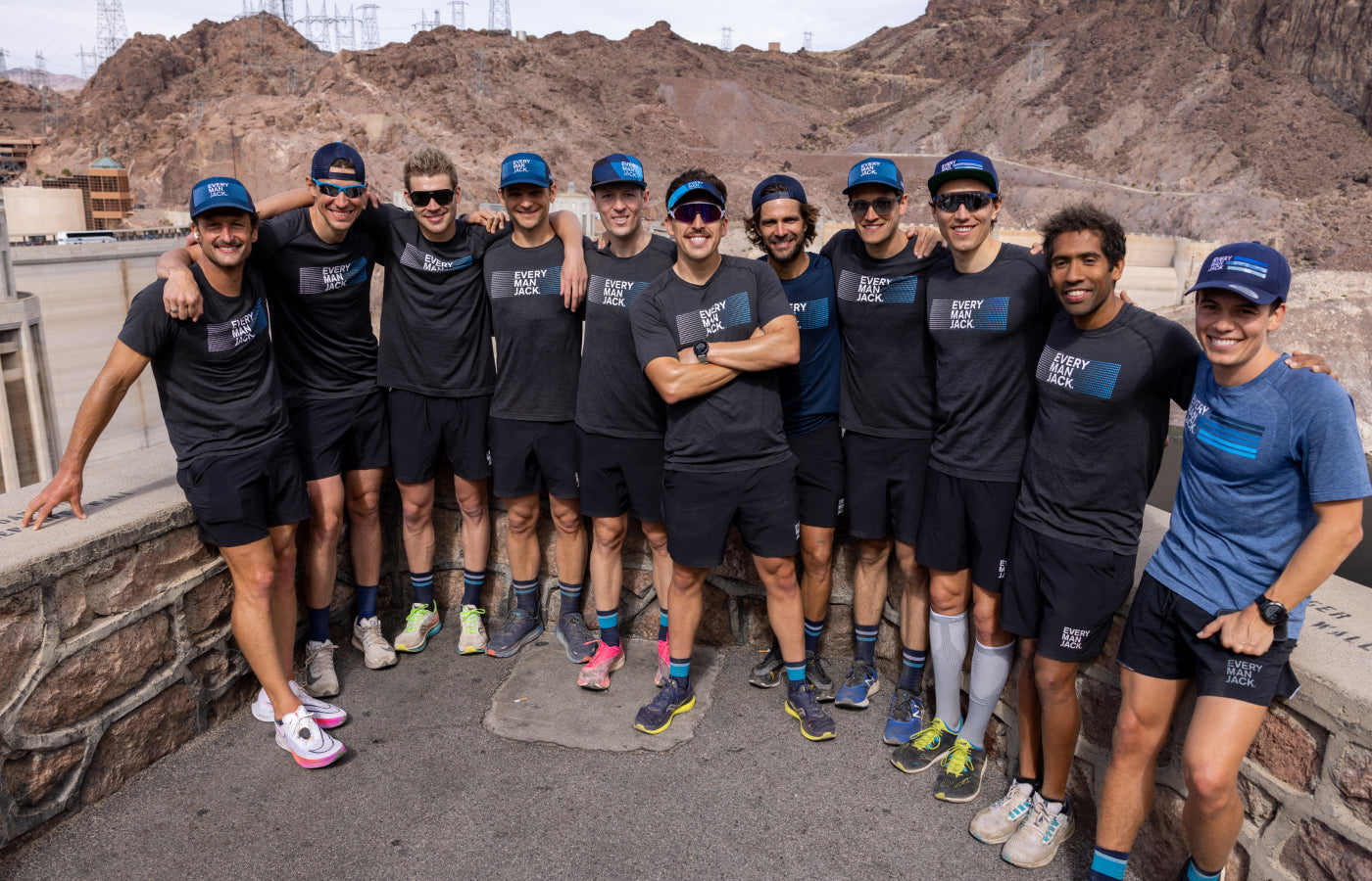 A group of 12 male athletes wearing matching Every Man Jack shirts and hats, posing together at a scenic location.