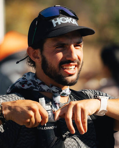 Man wearing a black cap with 'Hoka' branding, smiling outdoors.