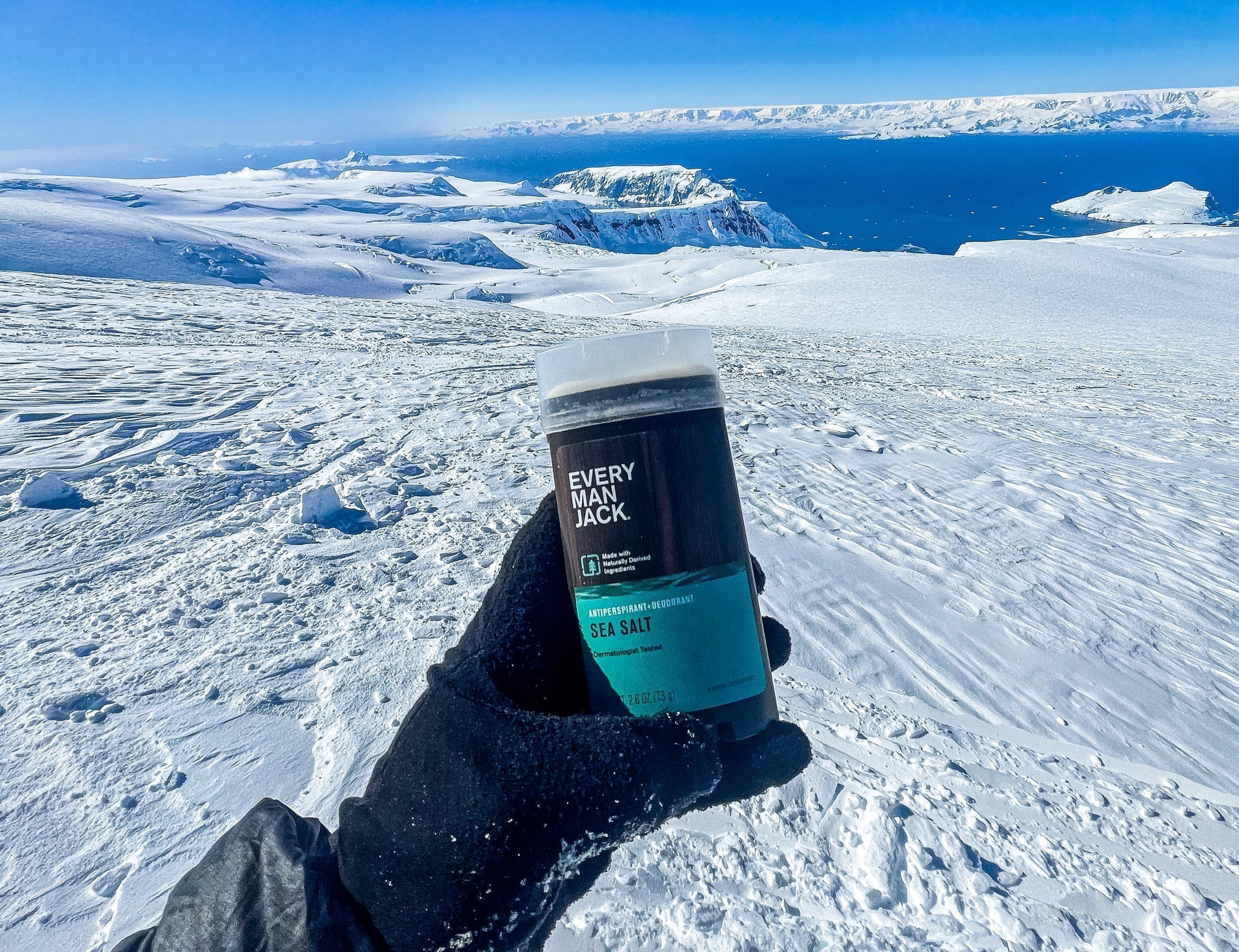 A person holding Every Man Jack Sea Salt Antiperspirant in a snowy landscape with mountains and ocean in the background.