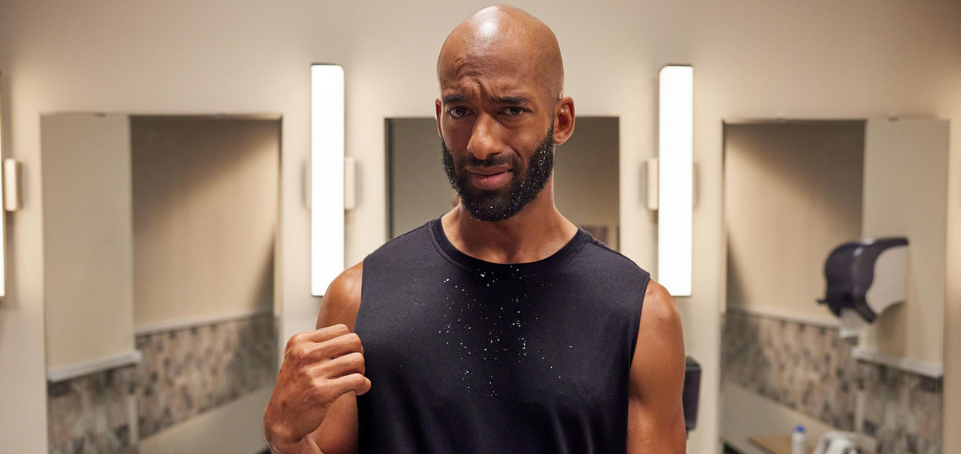 A man in a sleeveless shirt looking at the camera with a questioning expression in a bathroom setting.