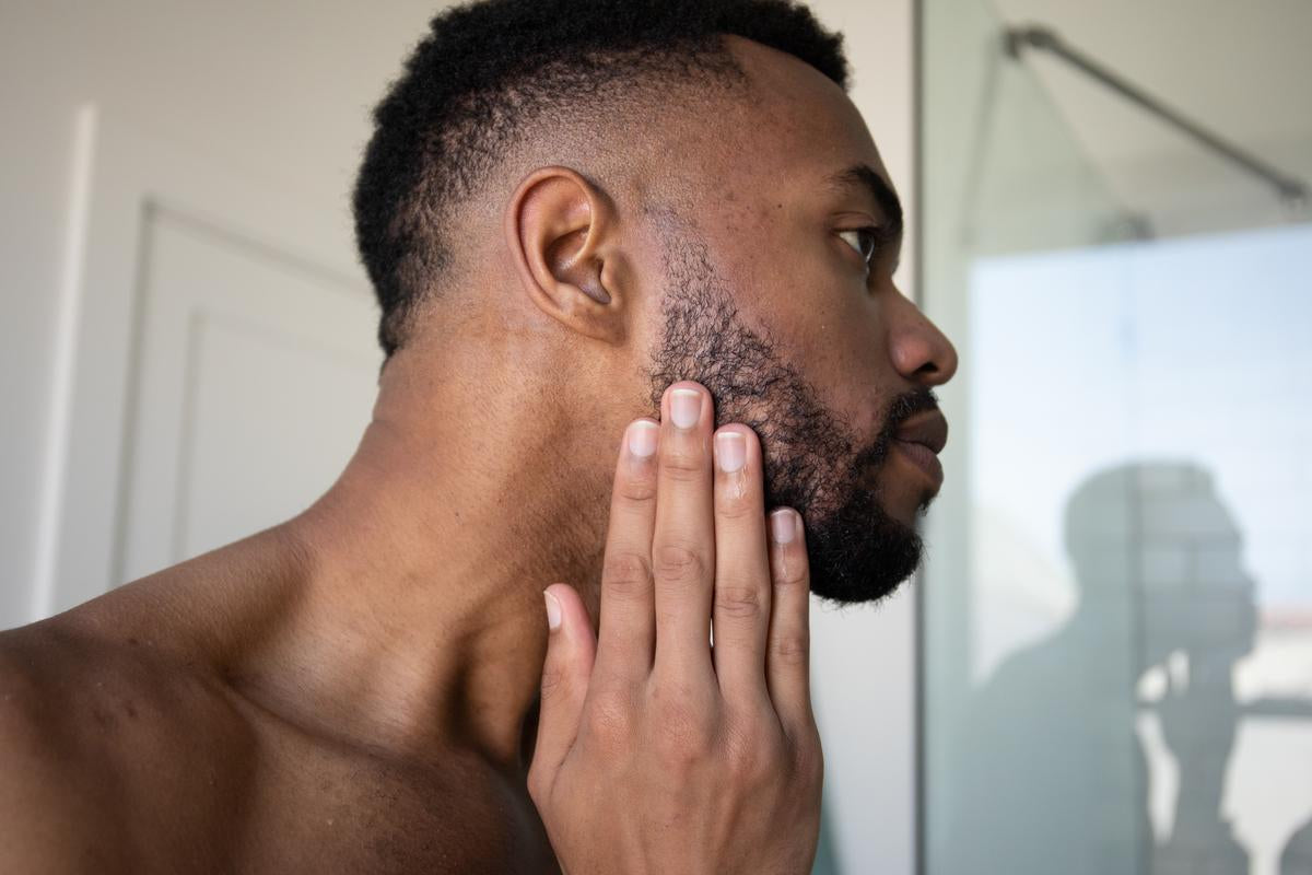 A man touching his beard while looking in a bathroom mirror.