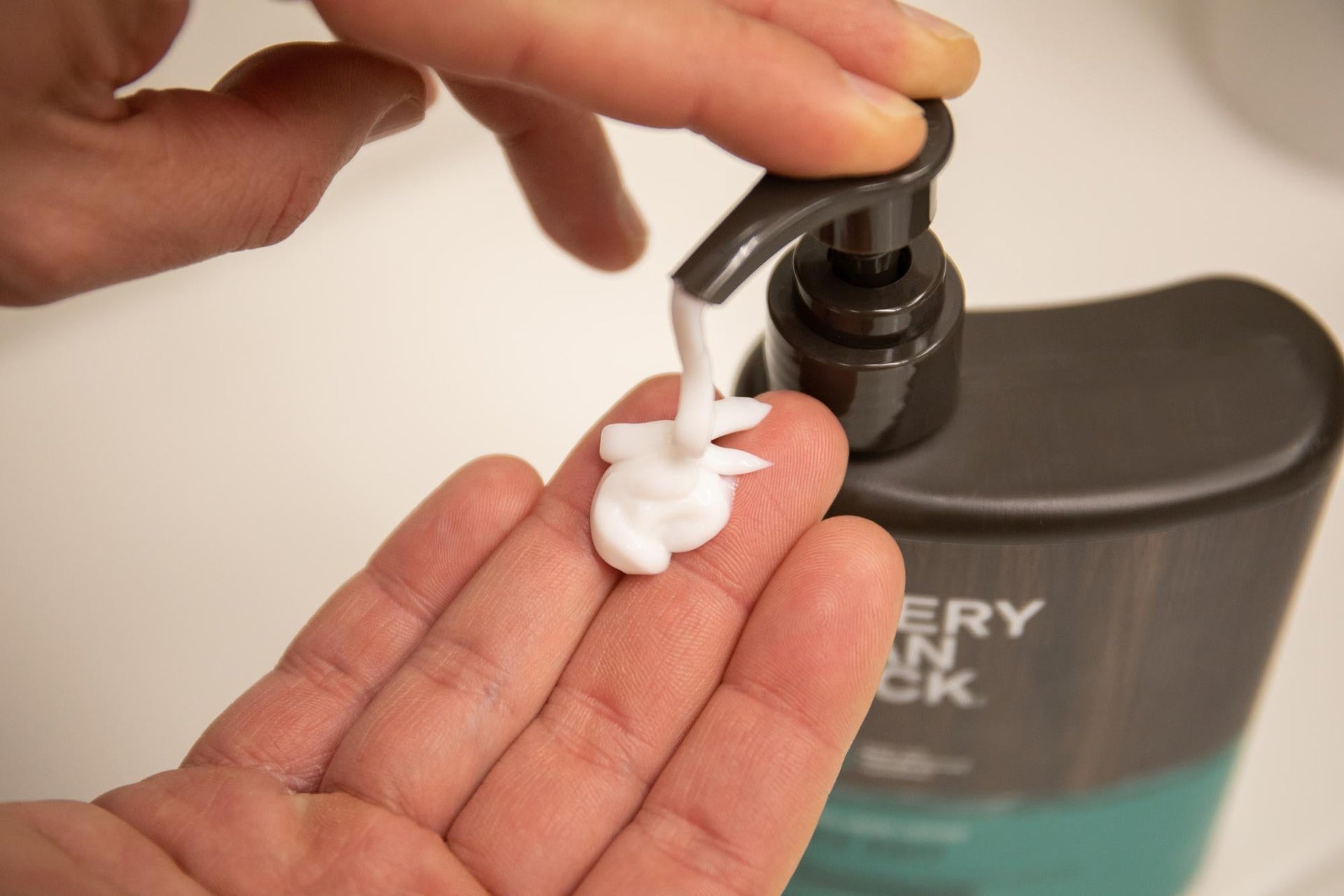 Hand dispensing white moisturizer from Every Man Jack pump bottle against a neutral background