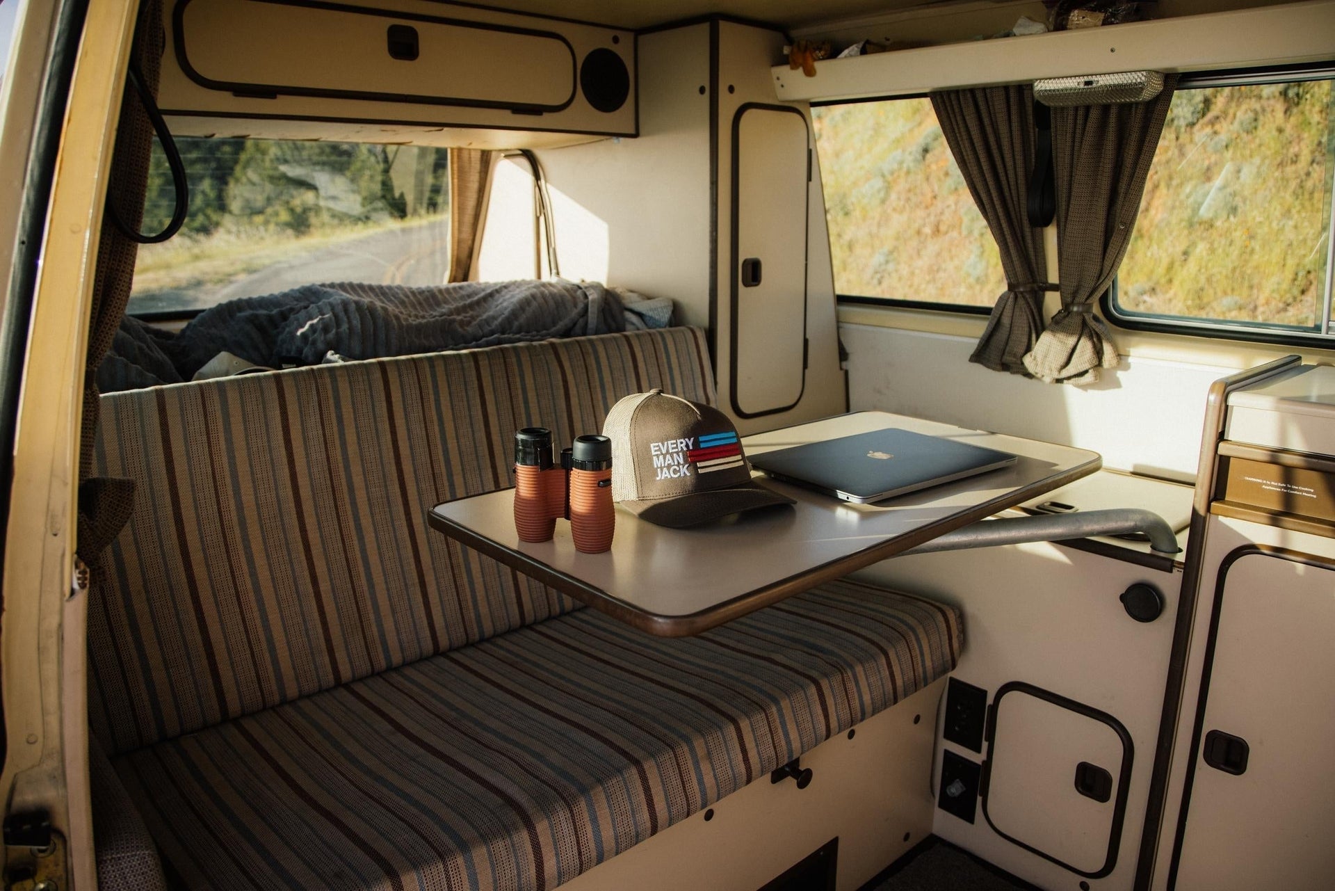 Every Man Jack cap and closed laptop on a camper van table with binoculars, striped bench seat, and sunlight streaming through the window