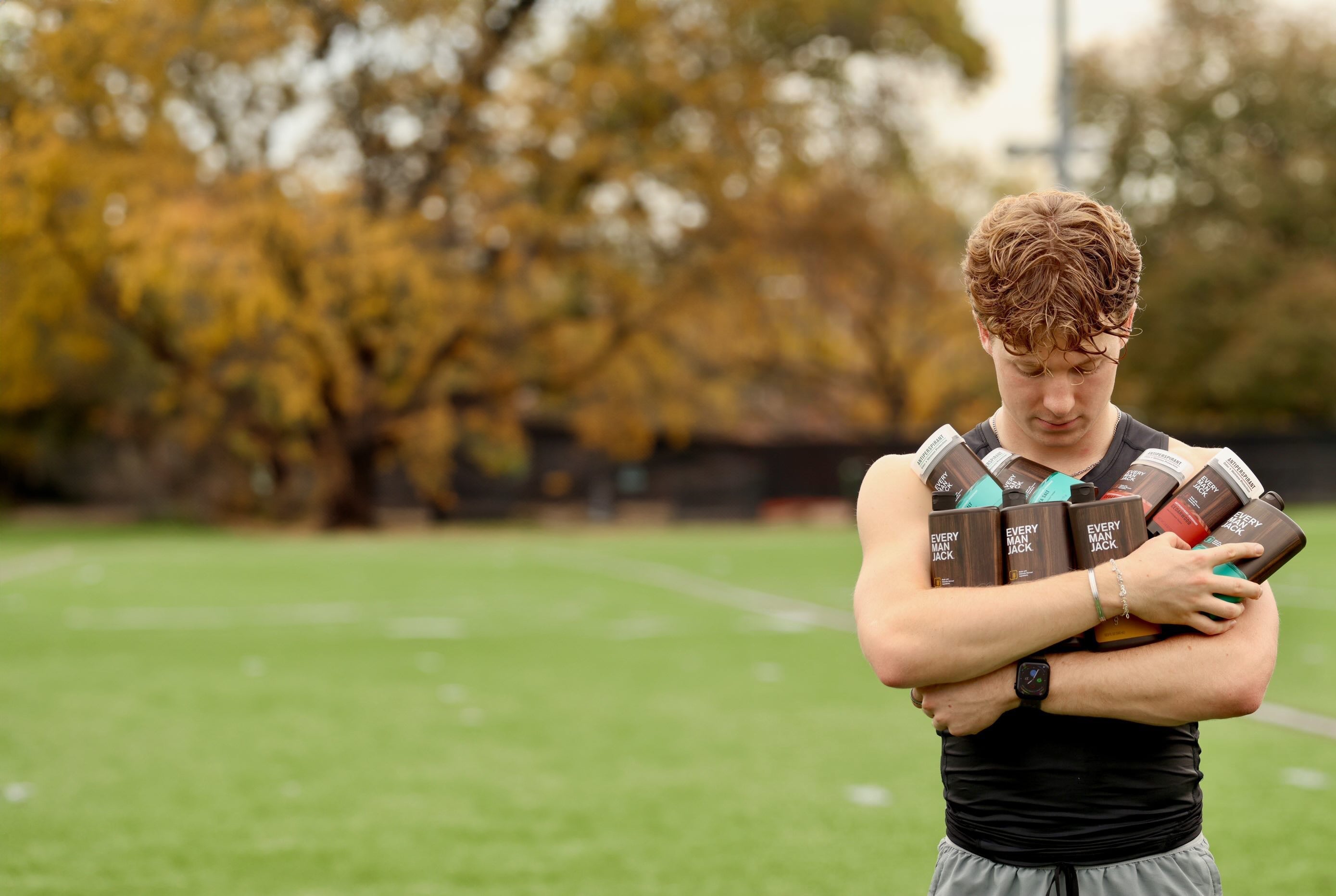 A young man holding several Every Man Jack grooming products on a field.