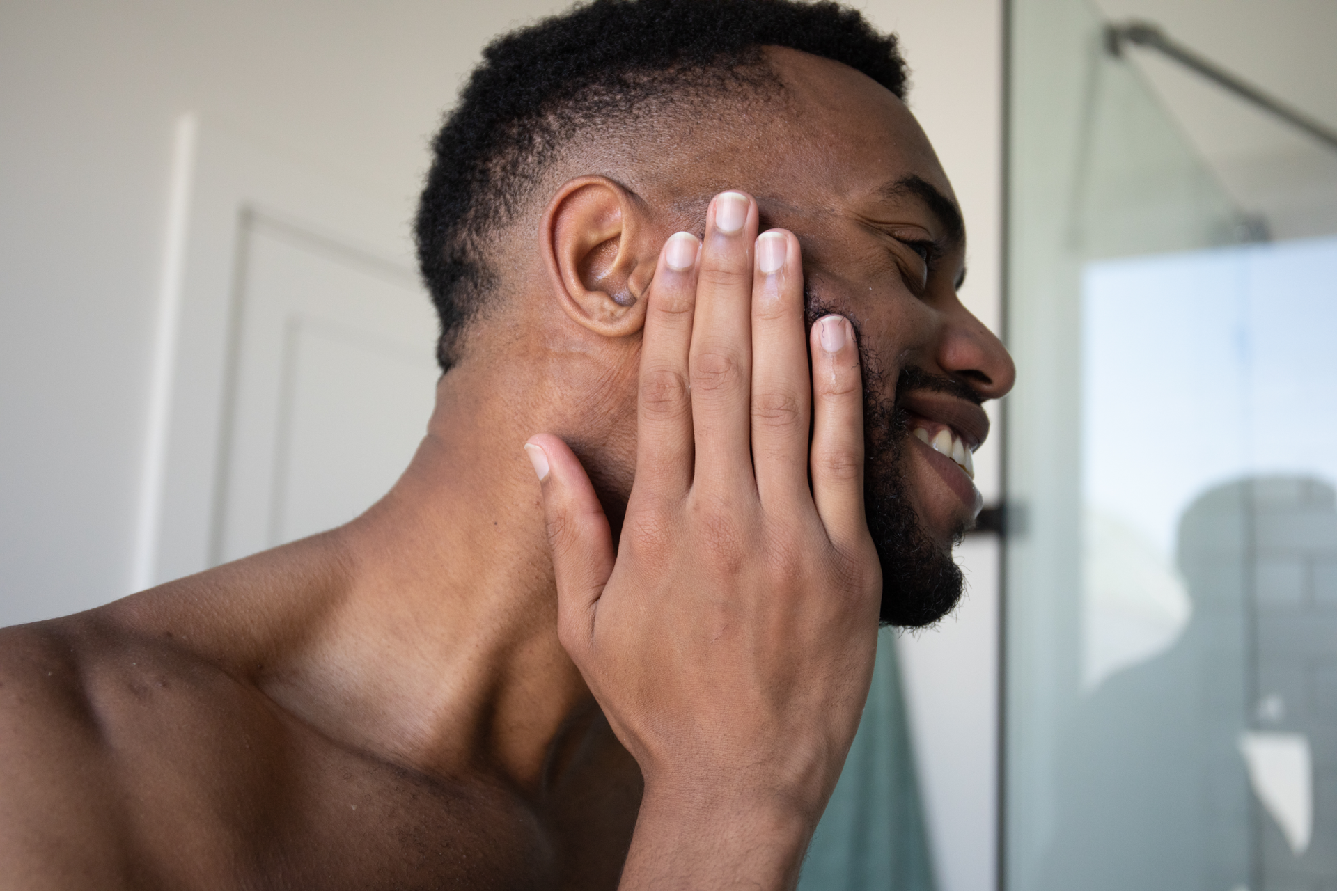 Smiling man pressing his hand to his cheek while grooming in a bright bathroom
