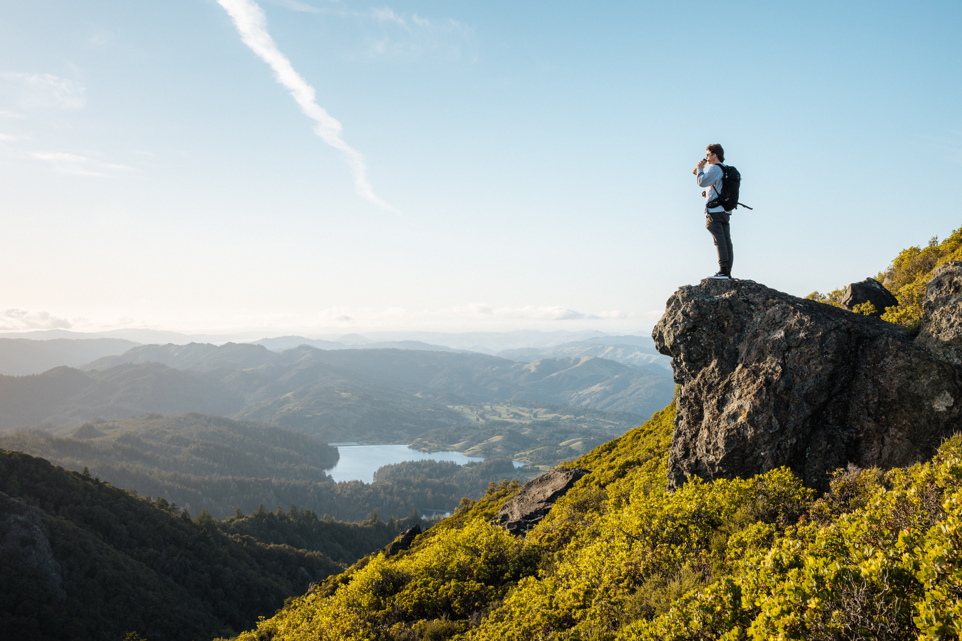A person standing on a rock overlooking a valley and lake under a clear sky.