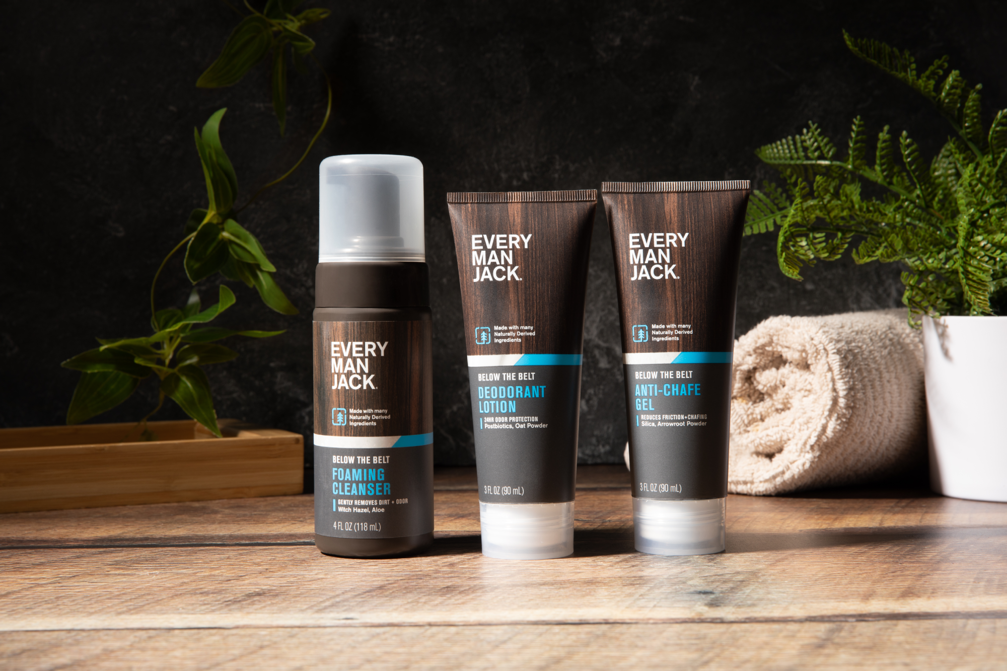 Three grooming products: Foaming Cleanser, Deodorant Lotion, and Anti-Chafe Gel by Every Man Jack on a wooden surface with a towel in the background.