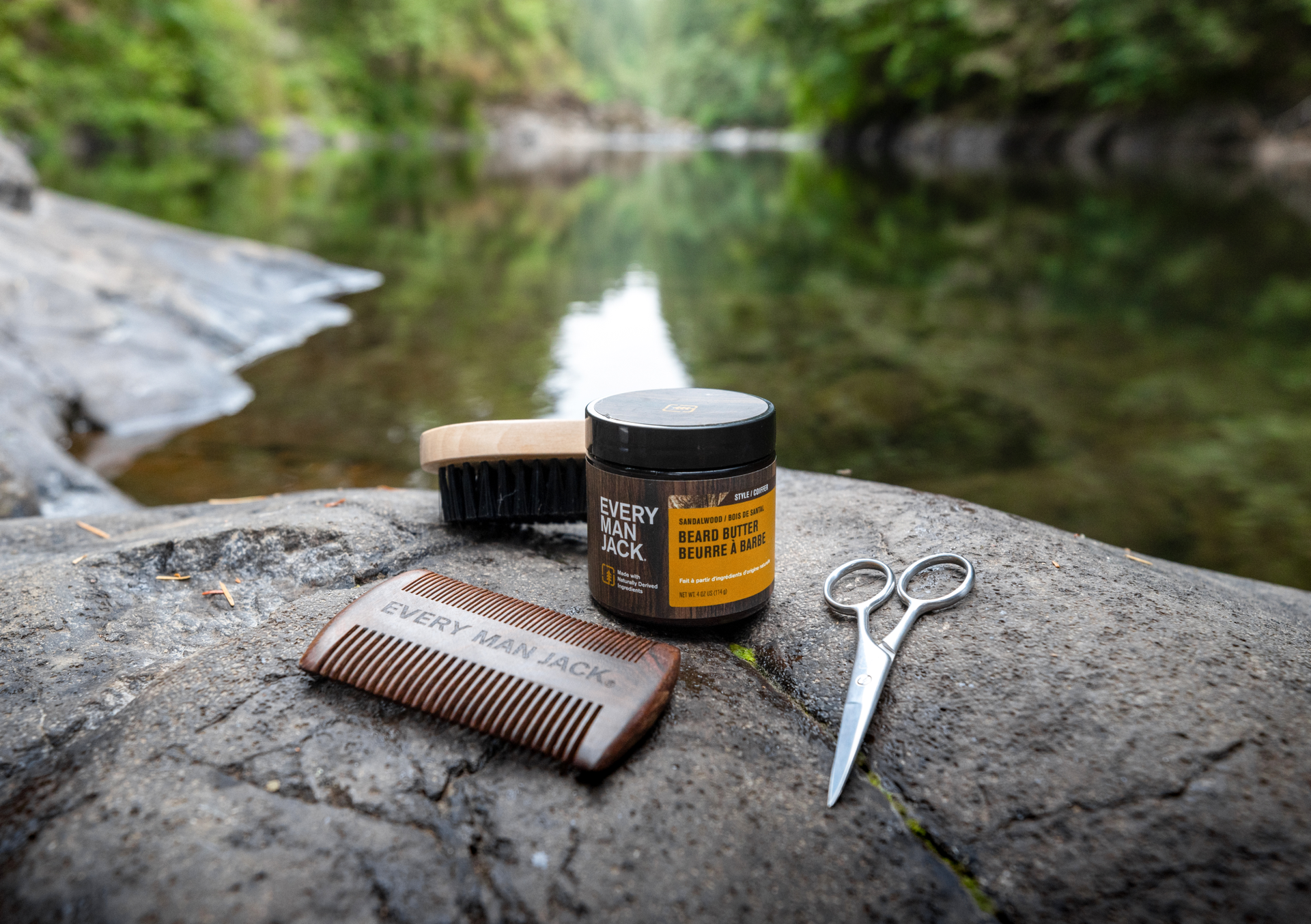 Beard grooming products including a comb, beard butter, and scissors on a rock by a river.