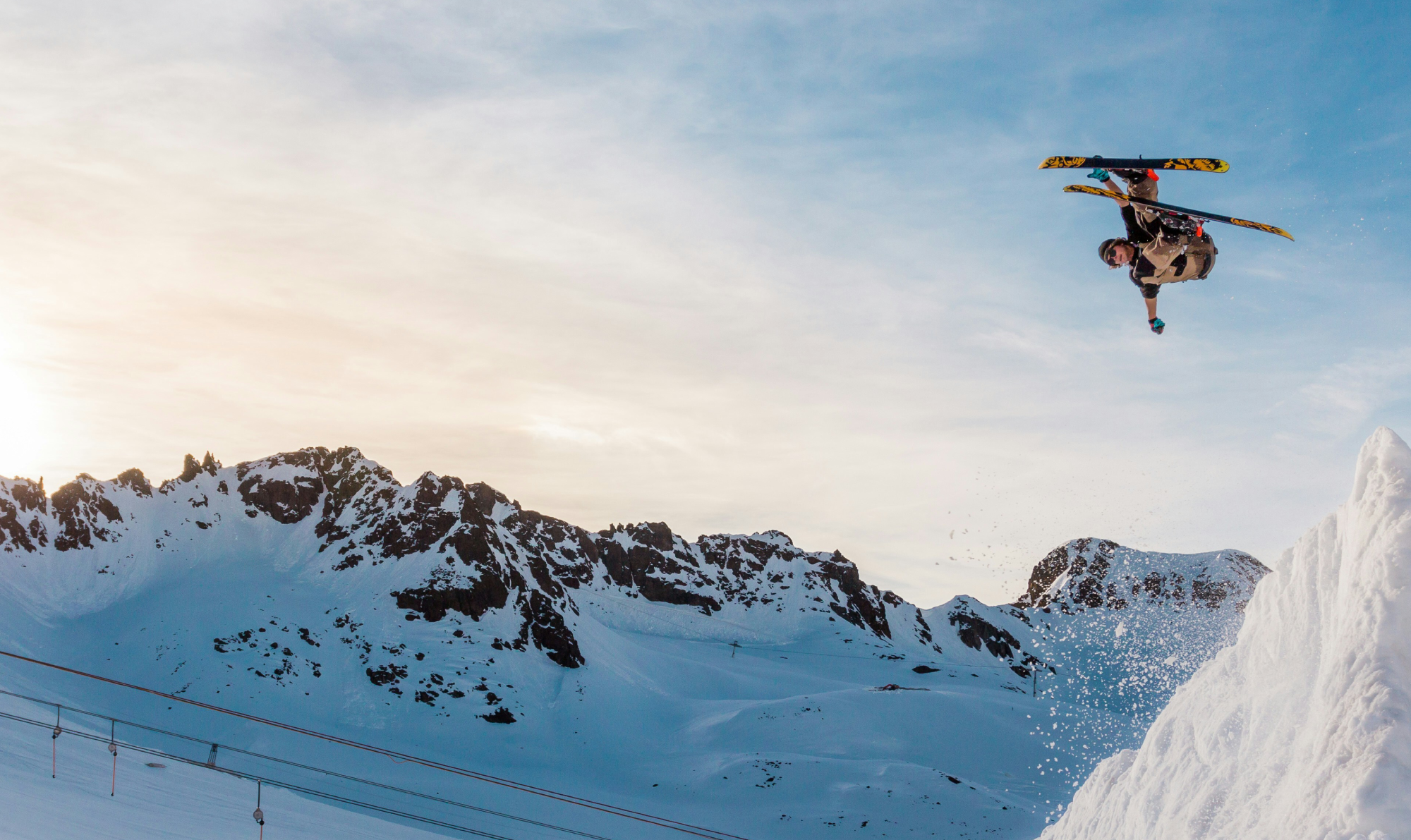 A skier performing a jump over a snow-covered slope with mountains in the background.