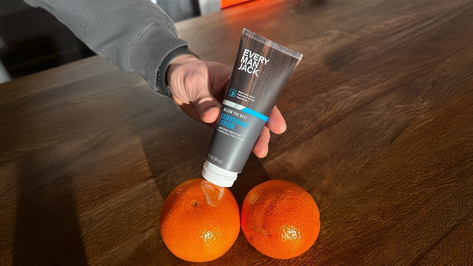 Handheld angled view of Every Man Jack Below the Belt deodorant lotion being squeezed onto two oranges on a wooden table