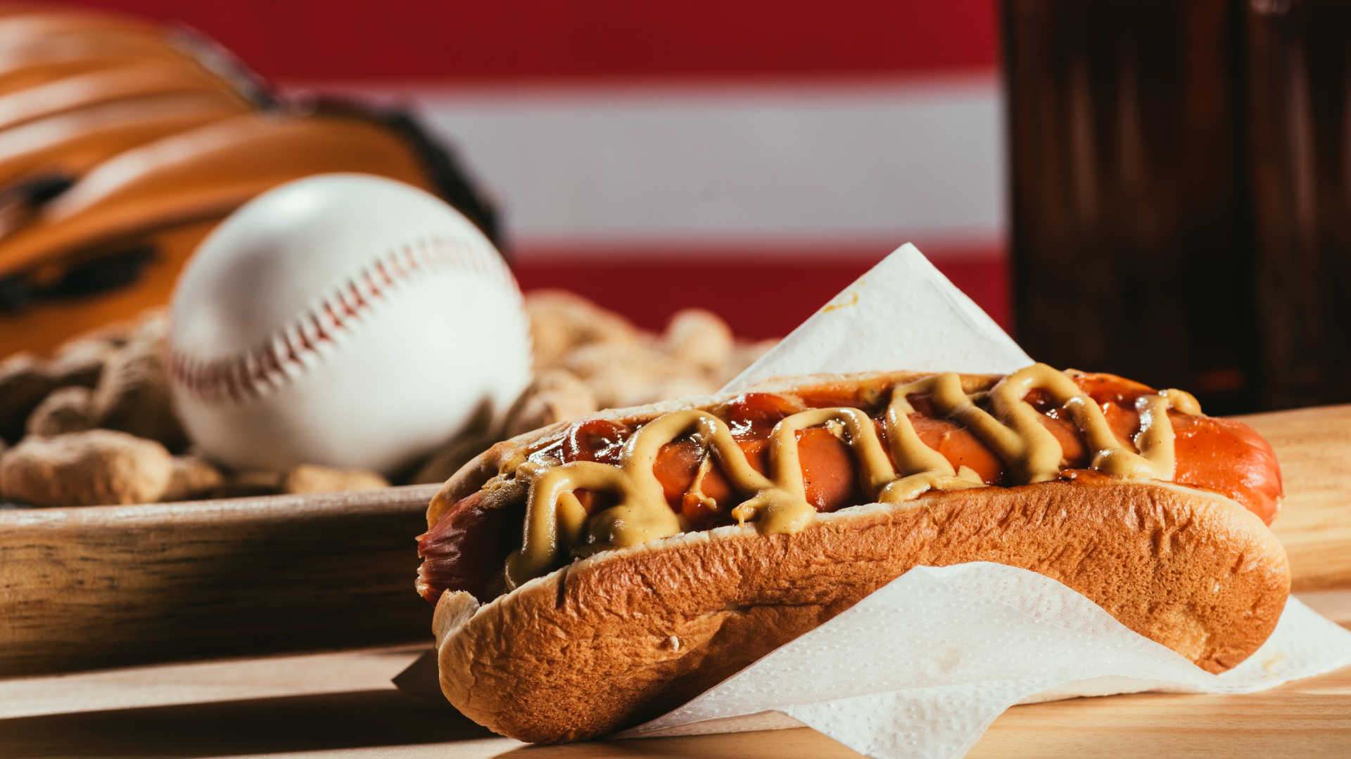 A hotdog with mustard on a bun, surrounded by a baseball, peanuts, and a drink.