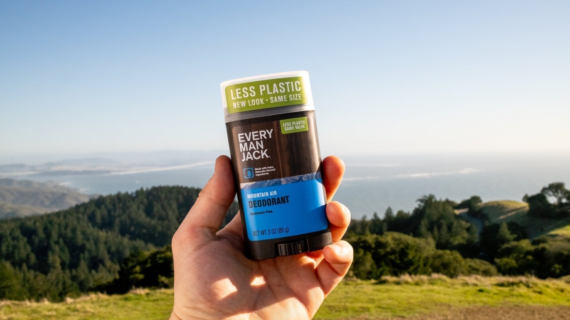 Close-up of Every Man Jack Mountain Air deodorant 3 oz showing Less Plastic cap and blue label, held before coastal mountains and ocean