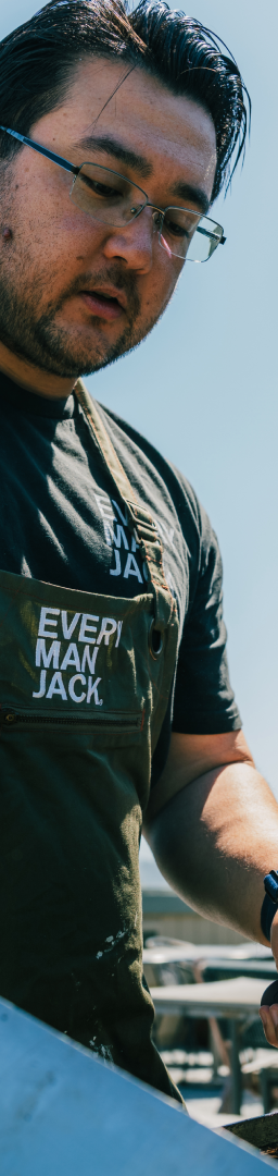 A person grilling outdoors, wearing an apron with 'Every Man Jack' logo.