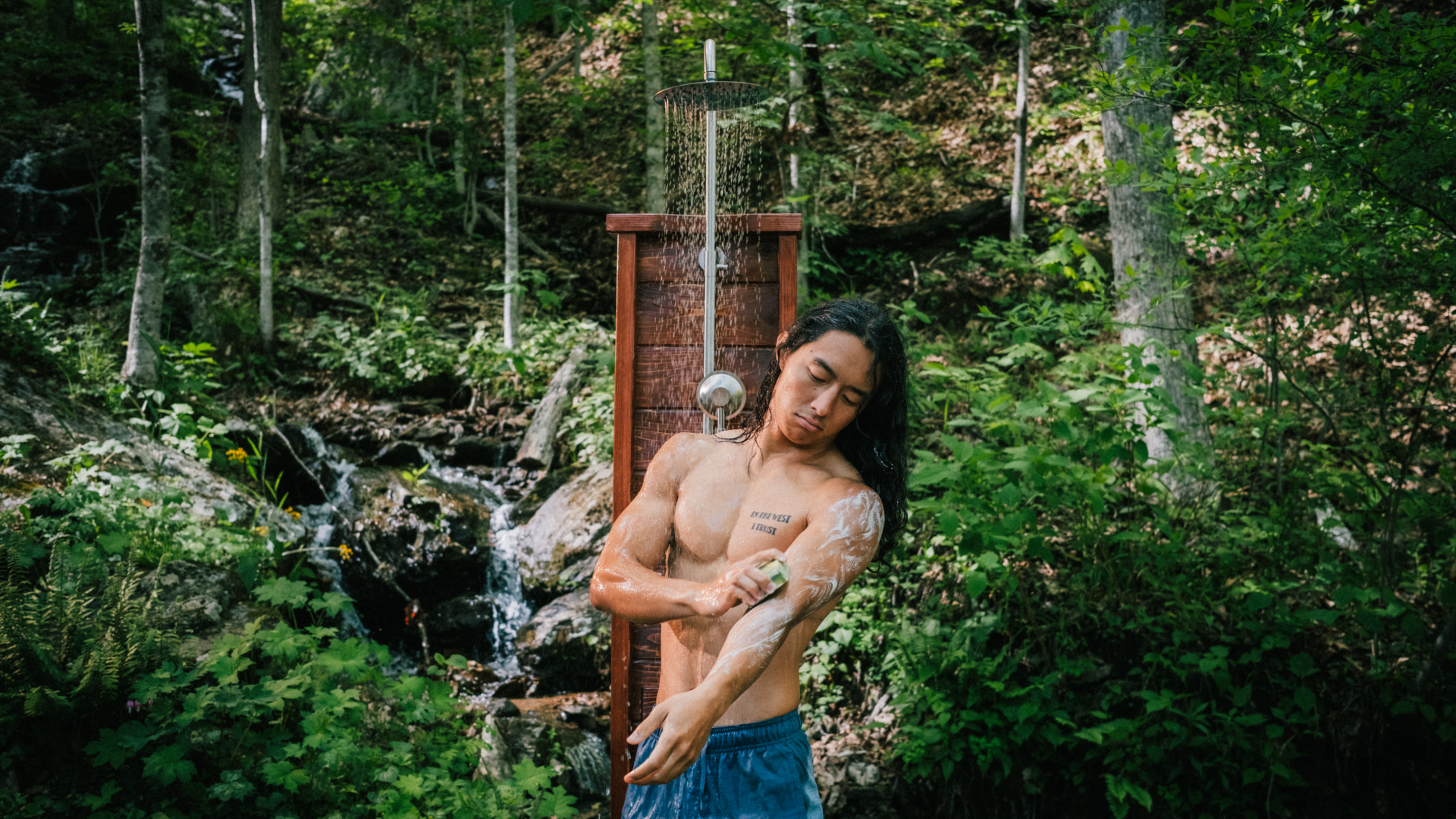 A person showering outdoors in a forest setting, applying soap to their arm.