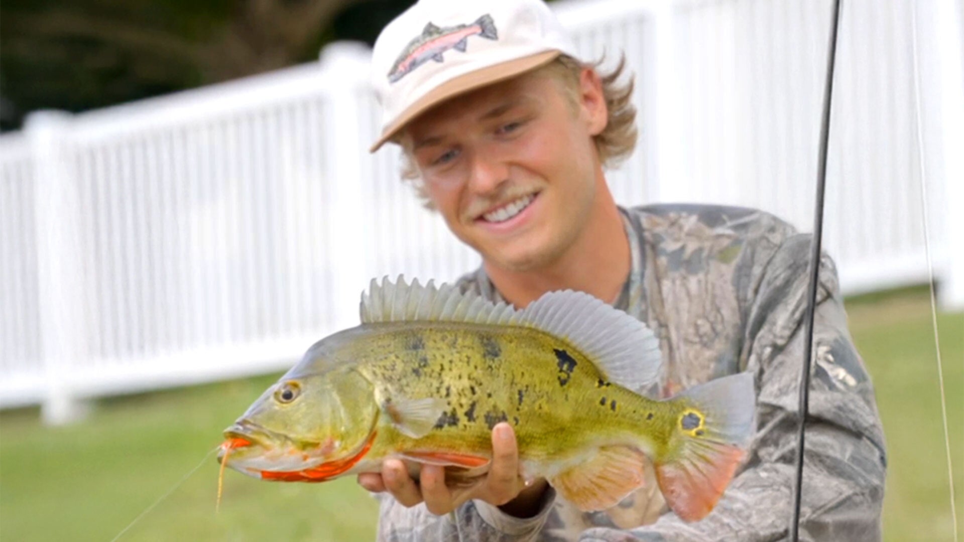 Young man wearing a cap and camouflage shirt holding a large fish with a fishing rod in the background outdoors
