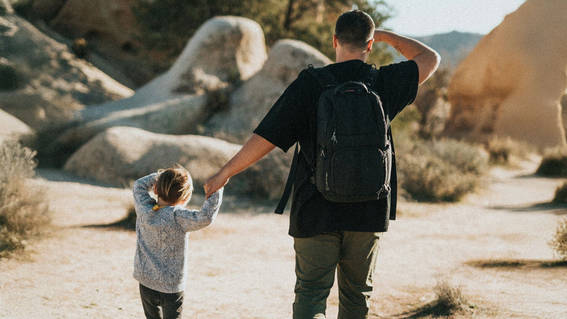 Man wearing a backpack holding hands with a young child walking on a sunlit rocky path outdoors.