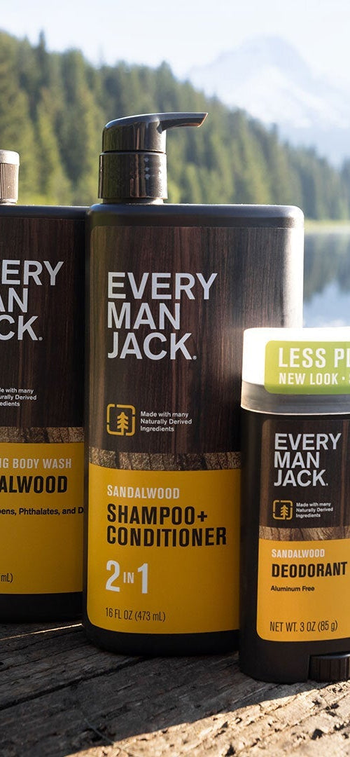 Three Every Man Jack sandalwood products: hydrating body wash, shampoo + conditioner 2 in 1, and deodorant with less plastic packaging, outdoors by a lake.
