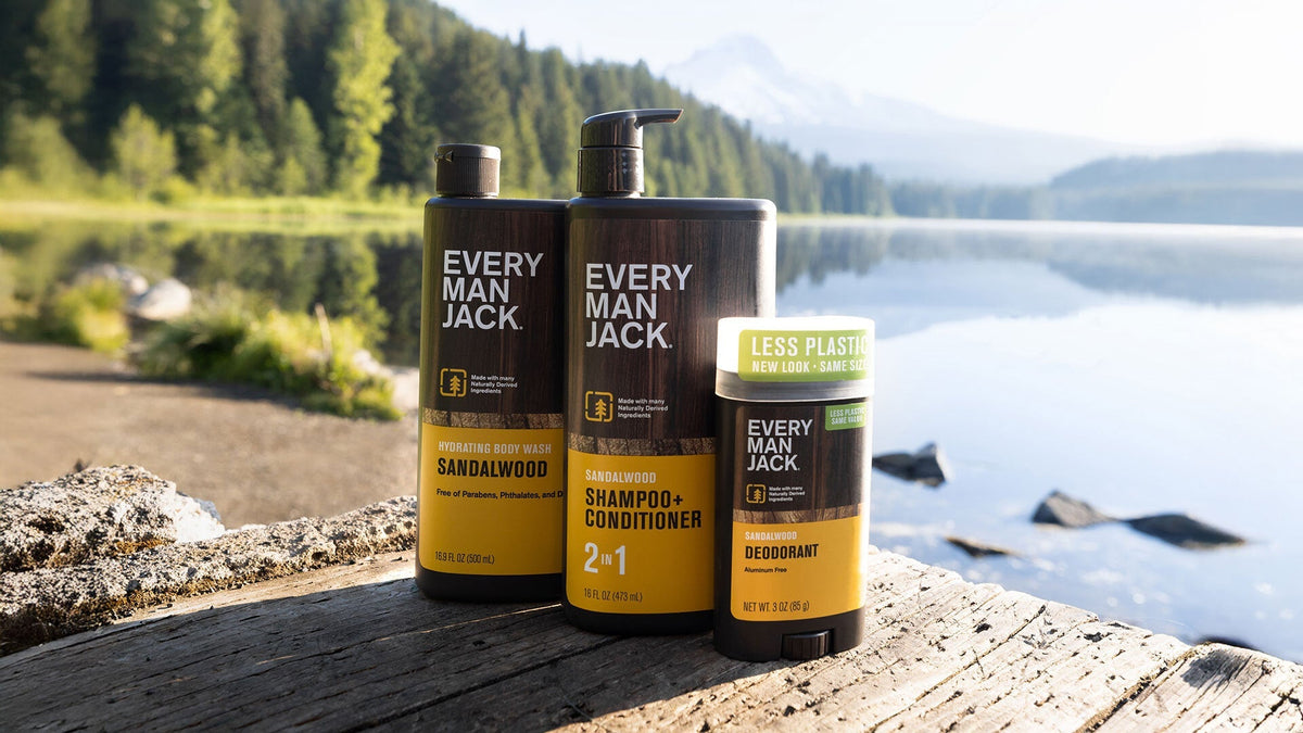 Three Every Man Jack sandalwood products: hydrating body wash, shampoo + conditioner 2 in 1, and deodorant with less plastic packaging, outdoors by a lake.
