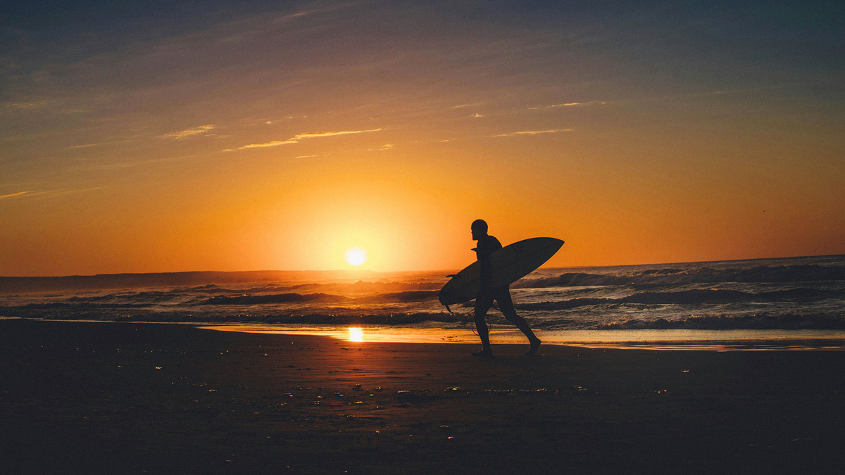 Surfer walking along a beach at sunset with warm orange sky and rolling waves