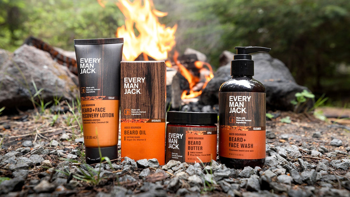 Every Man Jack Aged Bourbon grooming products arranged on rocks in front of a campfire in a forest setting