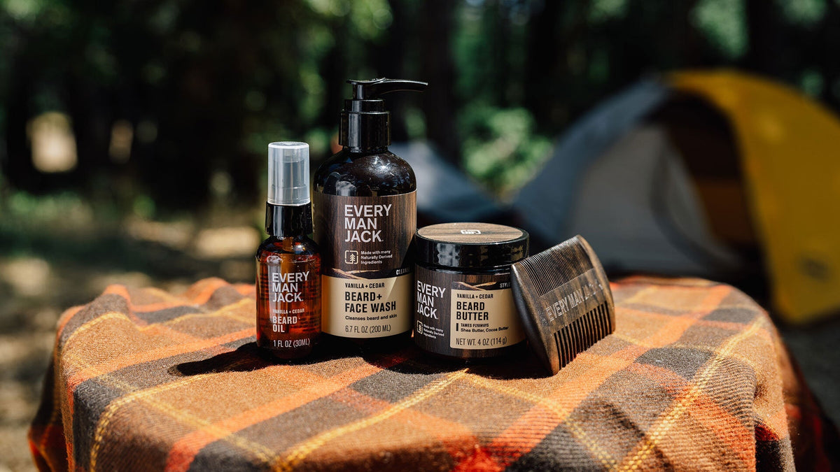 Front view of Vanilla and Cedar beard oil, Beard and Face Wash, beard butter, and comb on a plaid blanket with a tent in background