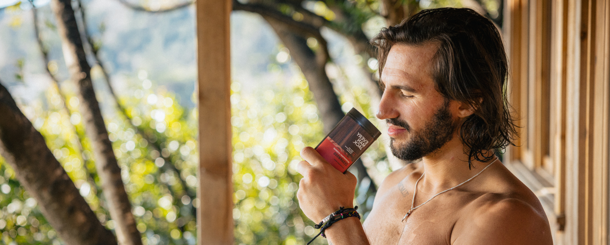 Shirtless man holding a bottle of Every Man Jack body wash on a sunlit wooden porch overlooking a forest