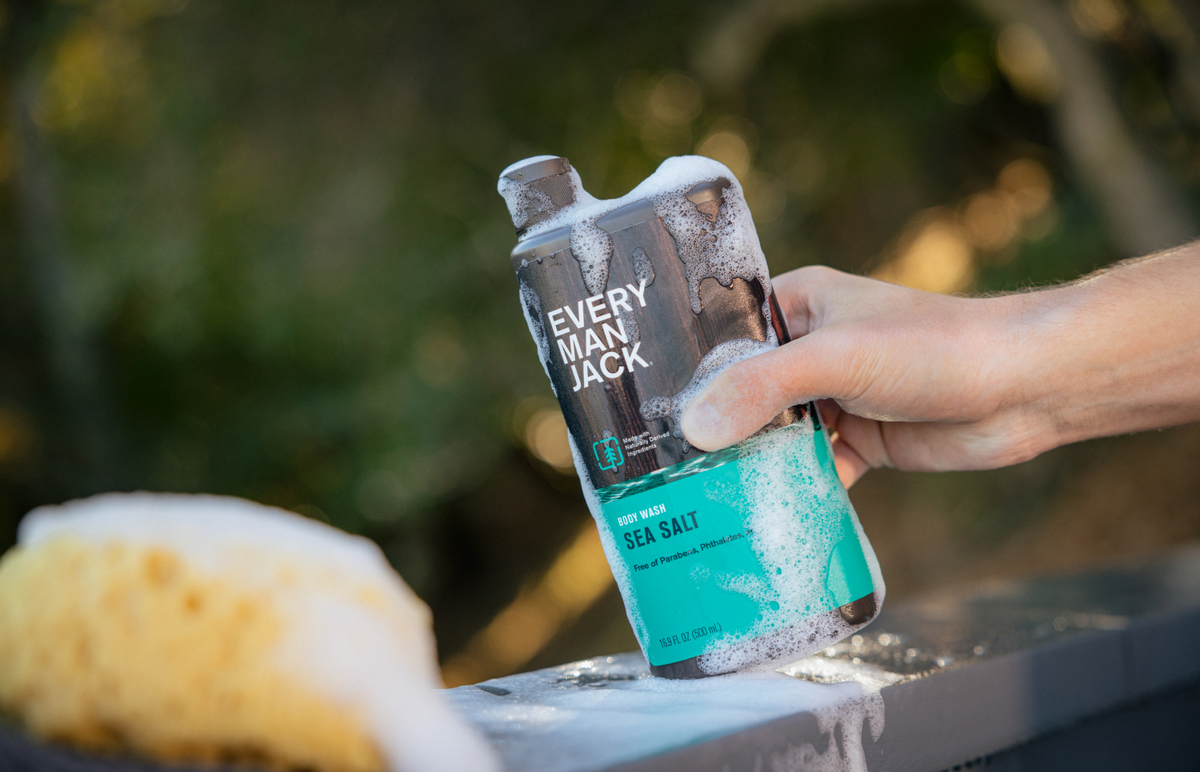 Angled view of Every Man Jack Sea Salt body wash bottle with soap suds, held by a hand against a blurred outdoor background