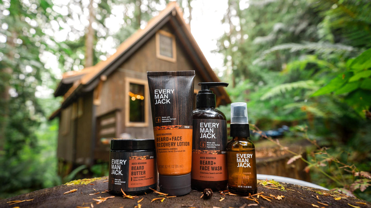 Front view of Aged Bourbon grooming set showcasing bottles, tube, and jar packaging with a rustic cabin and forest background